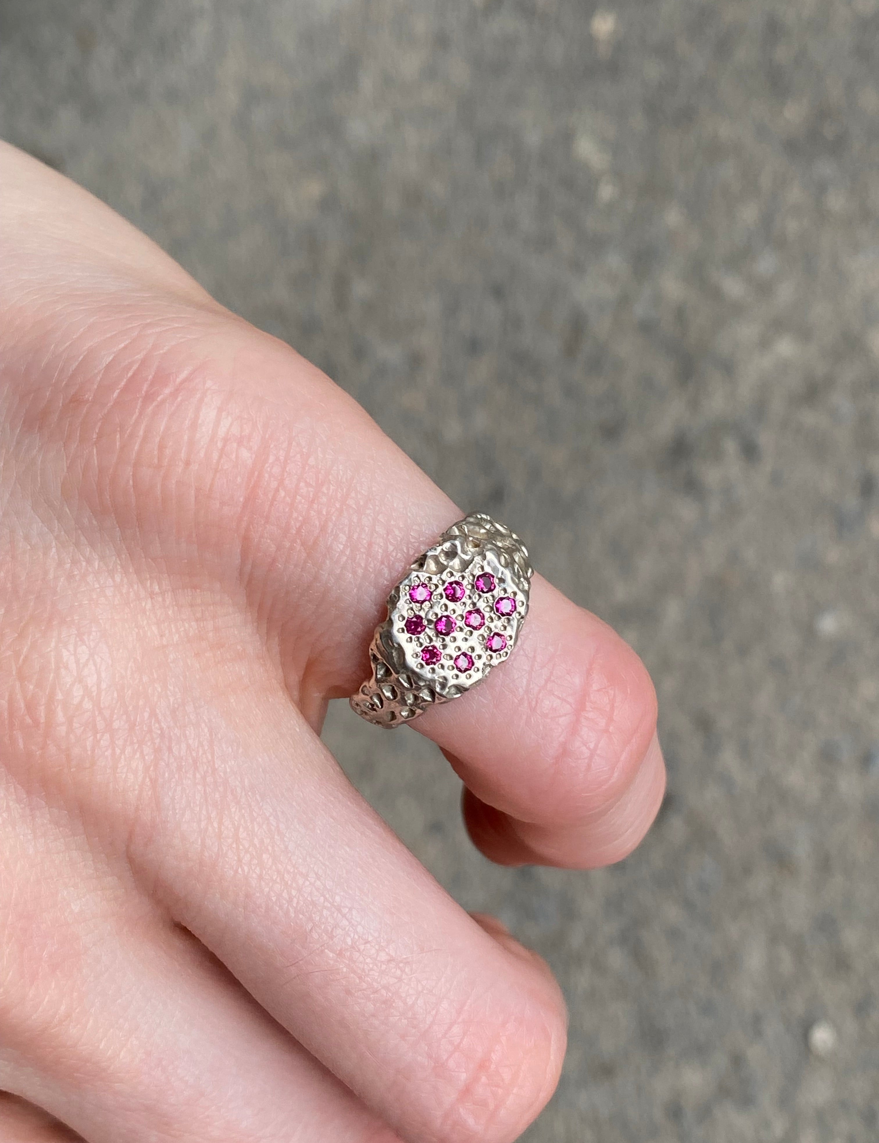 A silver ring with a cluster of small pink gemstones, worn on a person's finger against a gray concrete background.