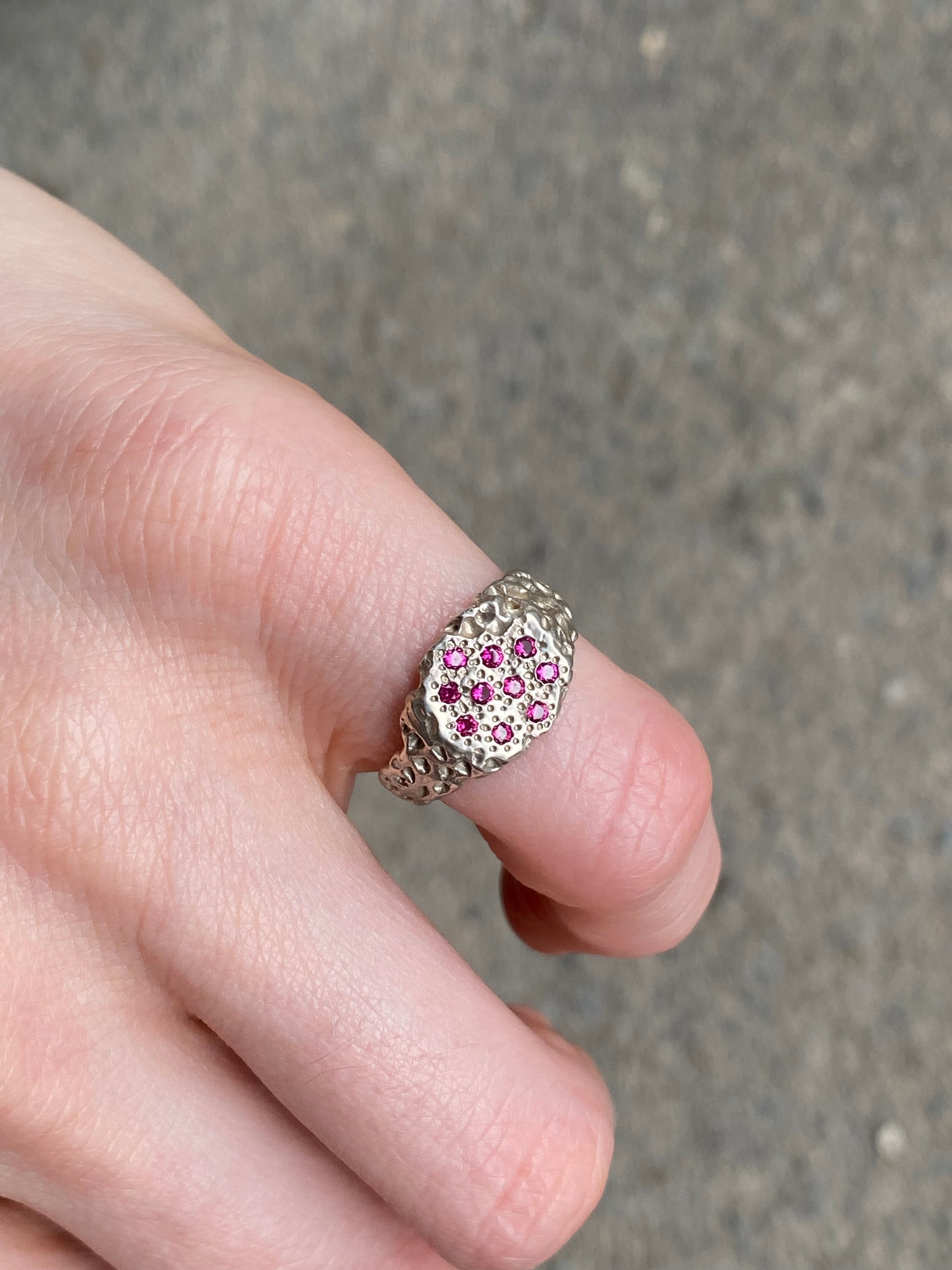 A silver ring with a cluster of small pink gemstones, worn on a person's finger against a gray concrete background.